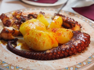 A plate of grilled octopus tentacles paired with large pieces of boiled potatoes, seasoned with red paprika or chili powder. The dish is presented on a decorated ceramic plate, suggesting a Mediterranean or Spanish style cuisine.