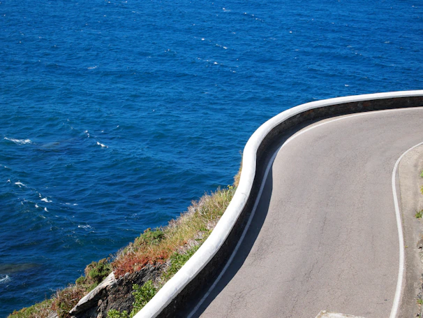 A winding coastal road viewed from above, bordered by deep blue ocean and beige cliffs.