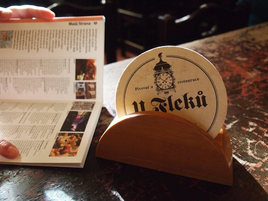 A close-up of a wooden coaster holder on a table, holding a round coaster with a vintage-style clock design and text in a foreign language. Next to it, a person is holding open a guidebook or brochure, showing text and images. The lighting casts a warm glow on the scene, highlighting the textures of the table and printed materials.