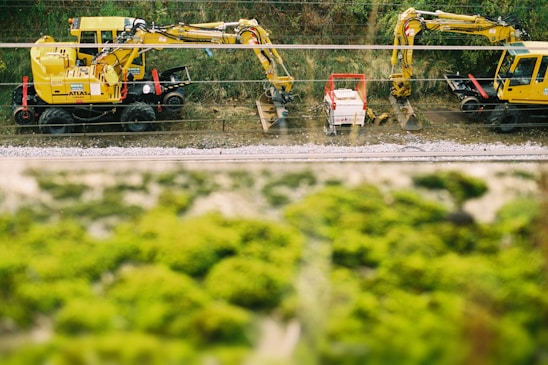 Two large yellow construction vehicles, possibly excavators, are positioned near a railway line. They are surrounded by greenery and some equipment between them, with tracks visible in the foreground.