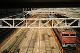 A train station with multiple railway tracks, featuring several trains at a halt under a large metal bridge. The tracks are surrounded by industrial structures, and there is a person working near one of the tracks wearing a high-visibility orange vest.