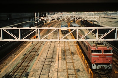 A train station with multiple railway tracks, featuring several trains at a halt under a large metal bridge. The tracks are surrounded by industrial structures, and there is a person working near one of the tracks wearing a high-visibility orange vest.