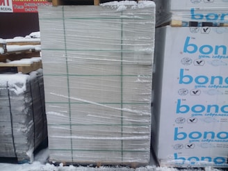 A large stack of construction materials, possibly insulation panels, is wrapped in plastic and secured with green straps. Snow covers the ground and some of the materials, indicating a cold outdoor environment. There are printed labels and logos in blue on the right side of some packages.