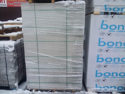 A large stack of construction materials, possibly insulation panels, is wrapped in plastic and secured with green straps. Snow covers the ground and some of the materials, indicating a cold outdoor environment. There are printed labels and logos in blue on the right side of some packages.