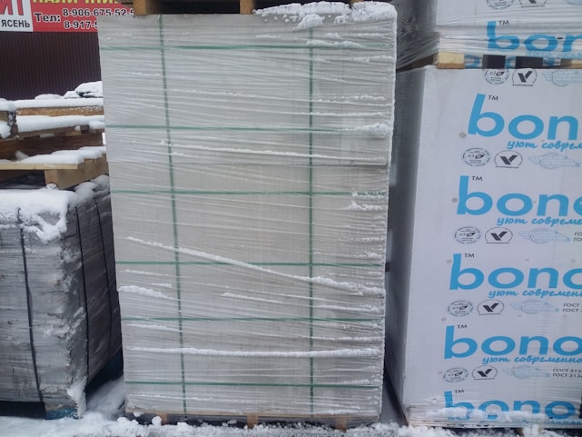 A large stack of construction materials, possibly insulation panels, is wrapped in plastic and secured with green straps. Snow covers the ground and some of the materials, indicating a cold outdoor environment. There are printed labels and logos in blue on the right side of some packages.