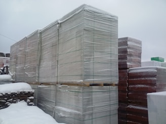 Large pallets covered in plastic wrap are stacked outdoors, all covered with a layer of snow. The scene is set against an overcast sky, suggesting a cold and snowy environment. The ground is not visible, likely under snow, and the pallets appear to be in an industrial or storage area.