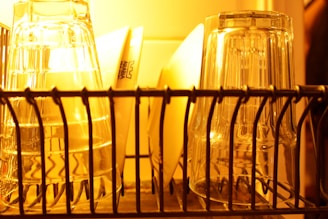 A close-up view of an organized dish rack with inverted glass cups and a few plates behind them. The lighting casts a warm, yellowish hue over the objects, creating reflections on the transparent surfaces of the glassware.