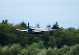 A modern training aircraft flying over a green military base.
