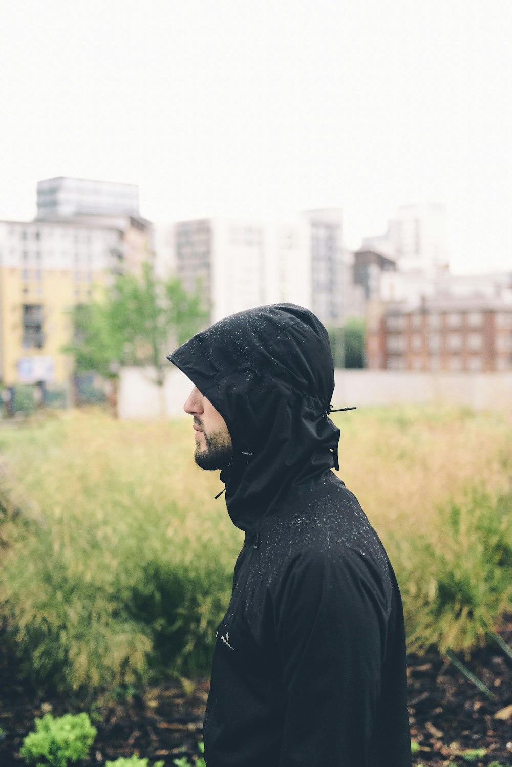 man wearing hoodie standing near grass with building background during daytime