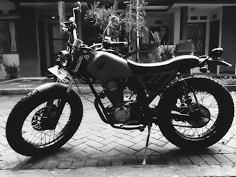 A classic motorcycle with a retro design, parked in front of a residential building. The vehicle features a large front headlamp, wire-spoke wheels, and a prominent engine block. The background reveals bricks and potted plants, creating a vintage atmosphere.