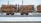 A train carrying large quantities of timber logs secured with orange straps on flatbed railcars. The scene takes place on a snowy railway track, suggesting cold weather conditions. The background shows industrial structures and electrical lines.