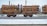 A train carrying large quantities of timber logs secured with orange straps on flatbed railcars. The scene takes place on a snowy railway track, suggesting cold weather conditions. The background shows industrial structures and electrical lines.