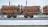 A train carrying large quantities of timber logs secured with orange straps on flatbed railcars. The scene takes place on a snowy railway track, suggesting cold weather conditions. The background shows industrial structures and electrical lines.