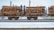 A train carrying large quantities of timber logs secured with orange straps on flatbed railcars. The scene takes place on a snowy railway track, suggesting cold weather conditions. The background shows industrial structures and electrical lines.