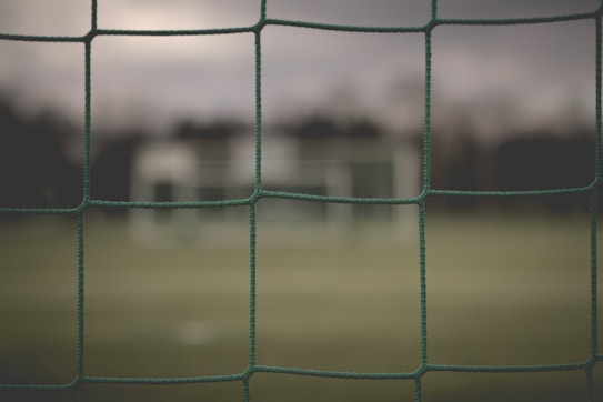 A close-up view of a green soccer net with blurred background showcasing a soccer goalpost and a grassy field. The focus is on the net, highlighting its texture and structure against the out-of-focus elements behind it, creating a serene, slightly melancholic ambiance.