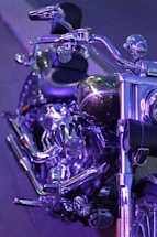 A close-up view of a motorcycle with shiny chrome detailing and purple lighting reflecting off its surfaces. The handlebars, seat, and parts of the engine are visible, showcasing the bike's polished metal and intricate design.
