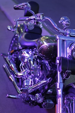 A close-up view of a motorcycle with shiny chrome detailing and purple lighting reflecting off its surfaces. The handlebars, seat, and parts of the engine are visible, showcasing the bike's polished metal and intricate design.