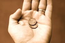A hand with a gold ring on the ring finger holds two small coins. The image has a sepia tone, creating a warm and vintage feel.