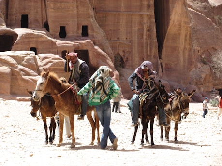 A group of people are riding donkeys in a desert landscape with rock formations in the background. Some of the riders are wearing traditional headscarves, and the scene suggests a rural or historical context.