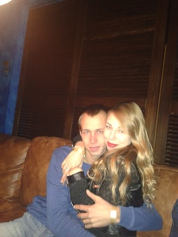 A couple is sitting close together on a brown leather couch. The man is wearing a blue long-sleeve shirt and has his arm around the woman, who has long blonde hair and is wearing a black jacket. The background includes dark wooden blinds and a blue wall on the left.