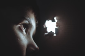 A person is peering through a small hole in a dark surface, with light illuminating the eye and the area around it. The image creates a sense of curiosity and mystery.