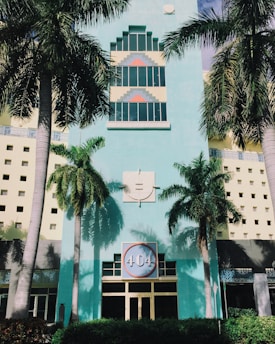 The image depicts an art deco style building with a pastel turquoise facade and geometric designs around the windows. Tall palm trees frame the building, casting distinctive shadows on the walls. A large circular sign with '404' is prominently displayed above the entrance, surrounded by architectural patterns.