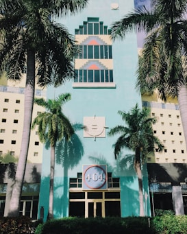 The image depicts an art deco style building with a pastel turquoise facade and geometric designs around the windows. Tall palm trees frame the building, casting distinctive shadows on the walls. A large circular sign with '404' is prominently displayed above the entrance, surrounded by architectural patterns.