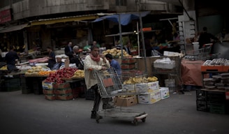 A bustling outdoor market scene with various stands and stalls displaying a wide array of fruits and vegetables. The foreground shows a man pushing a shopping cart filled with bananas and other goods, while other vendors and customers are seen in the background interacting and browsing the produce.