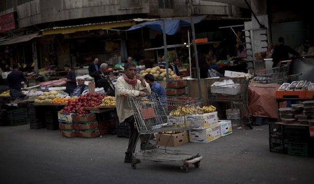 A bustling outdoor market scene with various stands and stalls displaying a wide array of fruits and vegetables. The foreground shows a man pushing a shopping cart filled with bananas and other goods, while other vendors and customers are seen in the background interacting and browsing the produce.