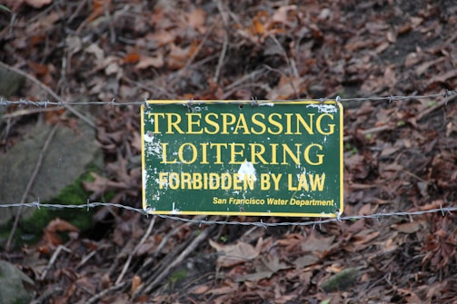A worn sign hangs on a barbed wire fence, reading 'TRESPASSING LOITERING FORBIDDEN BY LAW San Francisco Water Department'. The background consists of autumn leaves and underbrush, suggesting a natural or wooded area.
