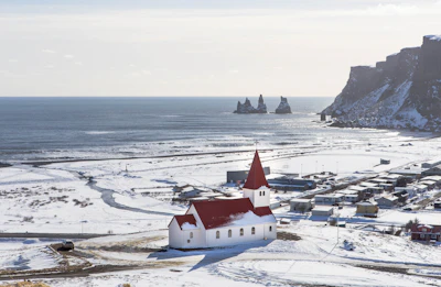 Vík’s church above the village in snow, with the Reynisdrangar stacks offshore — Iceland