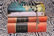 A stack of books on a patterned fabric background, topped with a string of black and white prayer beads. The titles include 'The Secret Lives of Baba Segi's Wives' and 'Half of a Yellow Sun', along with collections referred to as 'The African Trilogy' and 'The Cairo Trilogy'.