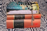 A stack of books on a patterned fabric background, topped with a string of black and white prayer beads. The titles include 'The Secret Lives of Baba Segi's Wives' and 'Half of a Yellow Sun', along with collections referred to as 'The African Trilogy' and 'The Cairo Trilogy'.