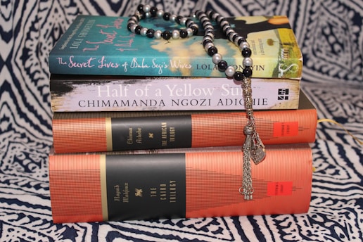 A stack of books on a patterned fabric background, topped with a string of black and white prayer beads. The titles include 'The Secret Lives of Baba Segi's Wives' and 'Half of a Yellow Sun', along with collections referred to as 'The African Trilogy' and 'The Cairo Trilogy'.