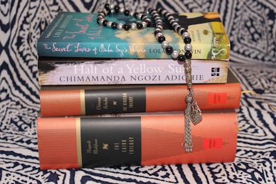 A stack of books on a patterned fabric background, topped with a string of black and white prayer beads. The titles include 'The Secret Lives of Baba Segi's Wives' and 'Half of a Yellow Sun', along with collections referred to as 'The African Trilogy' and 'The Cairo Trilogy'.