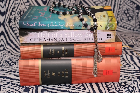 A stack of books on a patterned fabric background, topped with a string of black and white prayer beads. The titles include 'The Secret Lives of Baba Segi's Wives' and 'Half of a Yellow Sun', along with collections referred to as 'The African Trilogy' and 'The Cairo Trilogy'.
