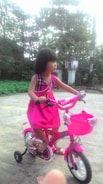 A young child wearing a bright pink dress is riding a small, colorful bicycle with training wheels. The bike features a matching pink basket at the front. The setting appears to be an outdoor area with greenery and a small structure in the background.
