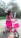 A young child wearing a bright pink dress is riding a small, colorful bicycle with training wheels. The bike features a matching pink basket at the front. The setting appears to be an outdoor area with greenery and a small structure in the background.
