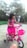 A young child wearing a bright pink dress is riding a small, colorful bicycle with training wheels. The bike features a matching pink basket at the front. The setting appears to be an outdoor area with greenery and a small structure in the background.