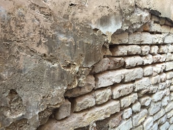 A weathered, partially plastered brick wall displaying signs of significant wear and deterioration. The plaster is chipped and flaking off, revealing the coarse and uneven brickwork underneath.