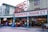 An entrance to a bustling public farmers market, marked by a large sign in red letters and a vintage clock above. The surrounding buildings are multi-story with various businesses, including a hotel. People are walking in and out of the market, some carrying bags, indicating active commerce. The atmosphere appears lively and urban.