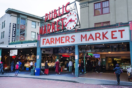 An entrance to a bustling public farmers market, marked by a large sign in red letters and a vintage clock above. The surrounding buildings are multi-story with various businesses, including a hotel. People are walking in and out of the market, some carrying bags, indicating active commerce. The atmosphere appears lively and urban.