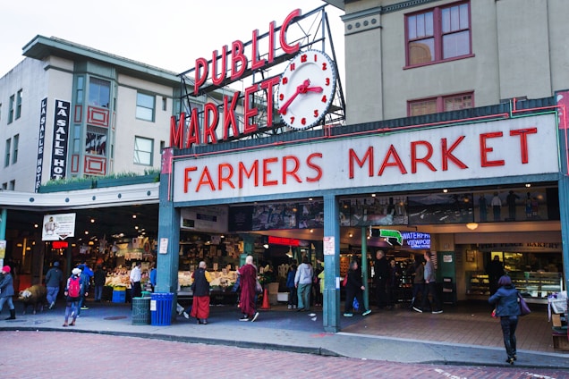 An entrance to a bustling public farmers market, marked by a large sign in red letters and a vintage clock above. The surrounding buildings are multi-story with various businesses, including a hotel. People are walking in and out of the market, some carrying bags, indicating active commerce. The atmosphere appears lively and urban.