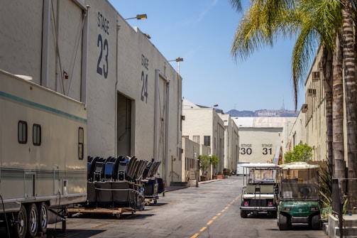 A studio backlot with large warehouse-like structures labeled as stages 23, 24, 30, and 31. There are trailers, equipment, and small utility vehicles parked along the road. Palm trees line one side, and the Hollywood sign is visible in the background.