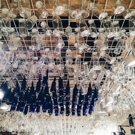 A ceiling installation composed of numerous clear and dark blue glass bottles hanging upside down from a metal grid. The bottles are arranged closely together in an artistic pattern.