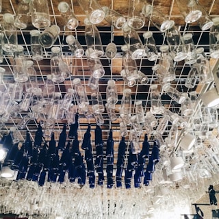 A ceiling installation composed of numerous clear and dark blue glass bottles hanging upside down from a metal grid. The bottles are arranged closely together in an artistic pattern.