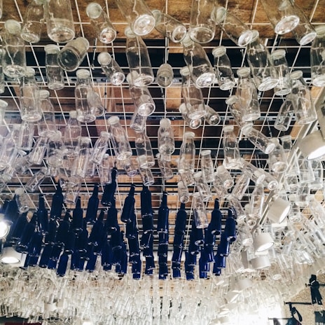 A ceiling installation composed of numerous clear and dark blue glass bottles hanging upside down from a metal grid. The bottles are arranged closely together in an artistic pattern.