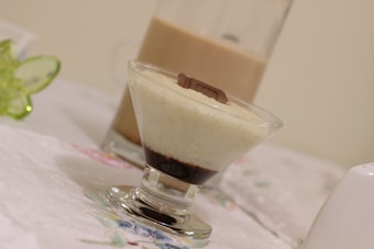 A dessert served in a clear glass with layers of creamy white and dark chocolate at the bottom, topped with a piece of milk chocolate. In the background, there is another glass filled with a light brown beverage.