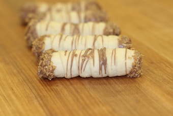 A series of rolled pastry cookies are lined up on a wooden surface. Each cookie is drizzled with chocolate and has ends coated in chopped nuts. The cookies are arranged in a diagonal line, creating a slightly blurred depth effect.