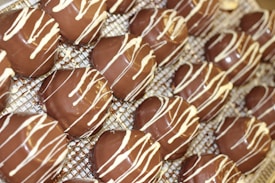 Several chocolate-covered treats are arranged on a metal cooling rack. Each treat is topped with a decorative drizzle of white chocolate. The smooth and shiny surface of the chocolate suggests they are freshly made.